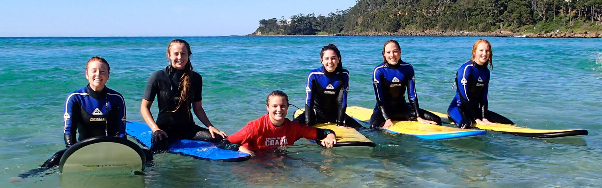 Surfing lessons girls in wetsuits sitting on surfboards in a row with instructor in the water in the middle