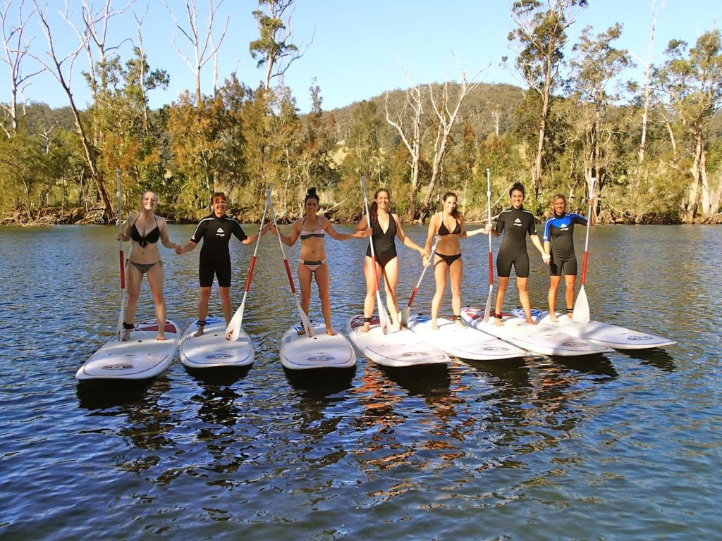 Smiling hen's party group posing and standing on paddle boards with bushy background