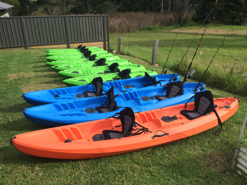 Green single kayaks, blue fishing kayaks and double orange kayak sitting on a row on green grass