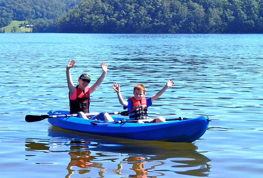 Smiling mother and son waving with life jackets on in blue kayak on the water