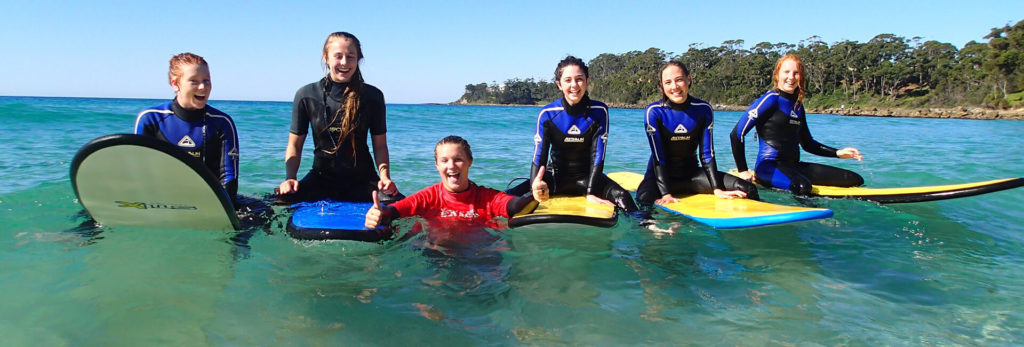 Wide shot of girls sitting on surfboards with a coach in the water