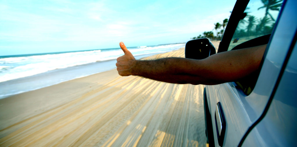 Arm outstretched out of car with thumbs up with beach in the background