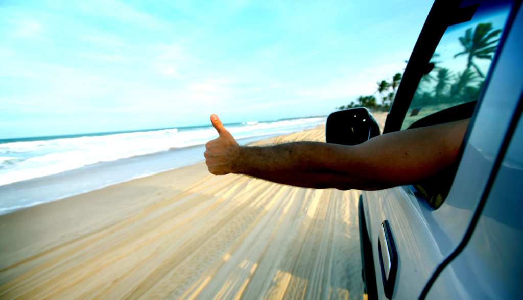 Arm outstretched out of car with thumbs up with beach in the background