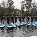 Group Posing For Photo Standing On Paddle Boards With Paddles In Their Hands On The Water