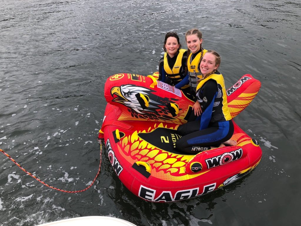 Three women with yellow life jackets seated on wake board on the water