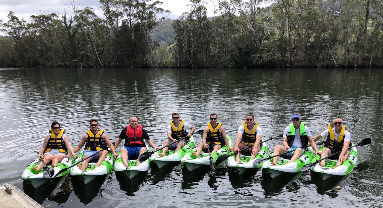 Large group of people with life jackets on seated in single kayaks in a row on the water with bush on the banks in the background