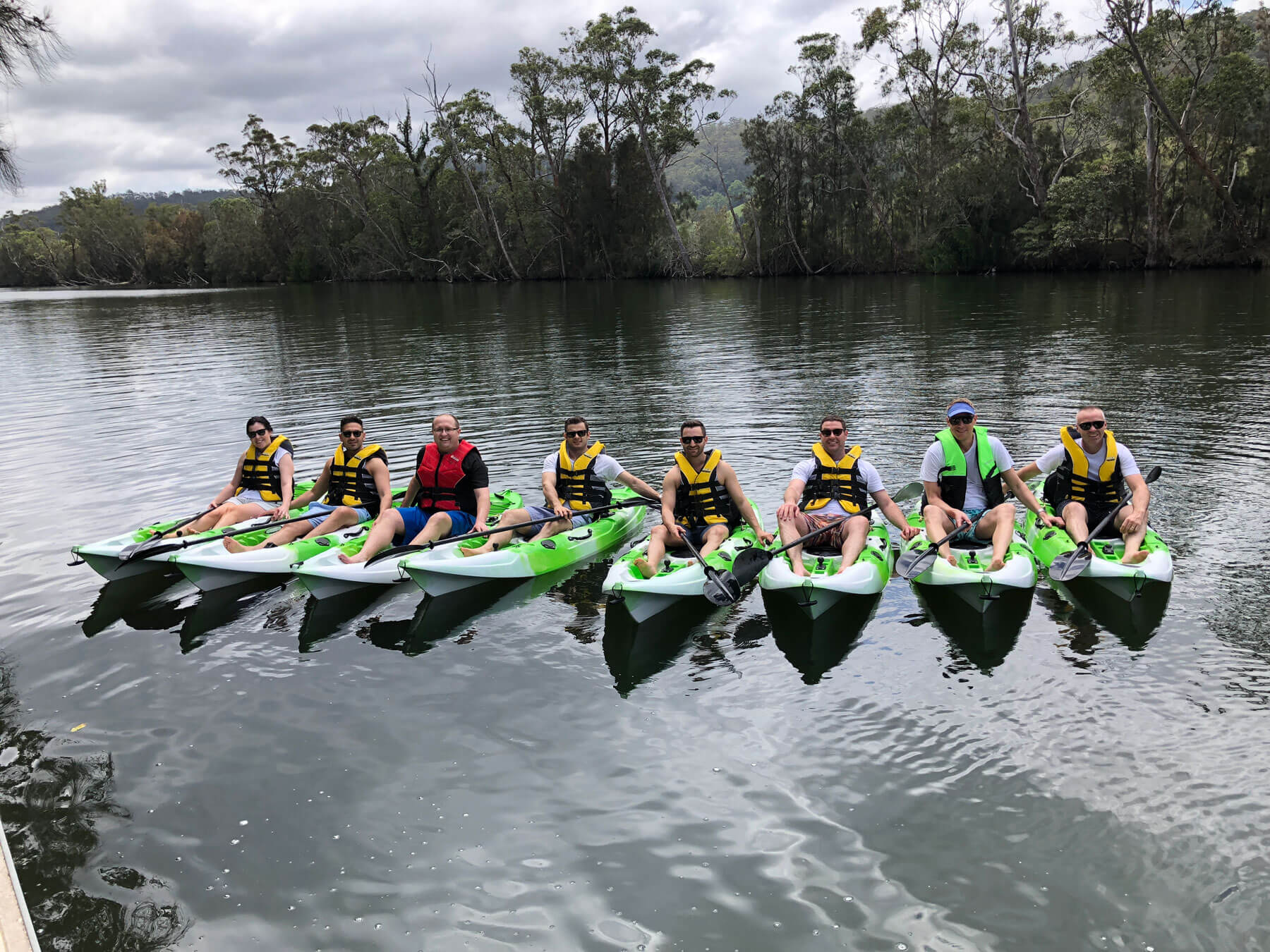 Group of people wearing life jackets seated in individual kayaks in a row on the water with bush in the background