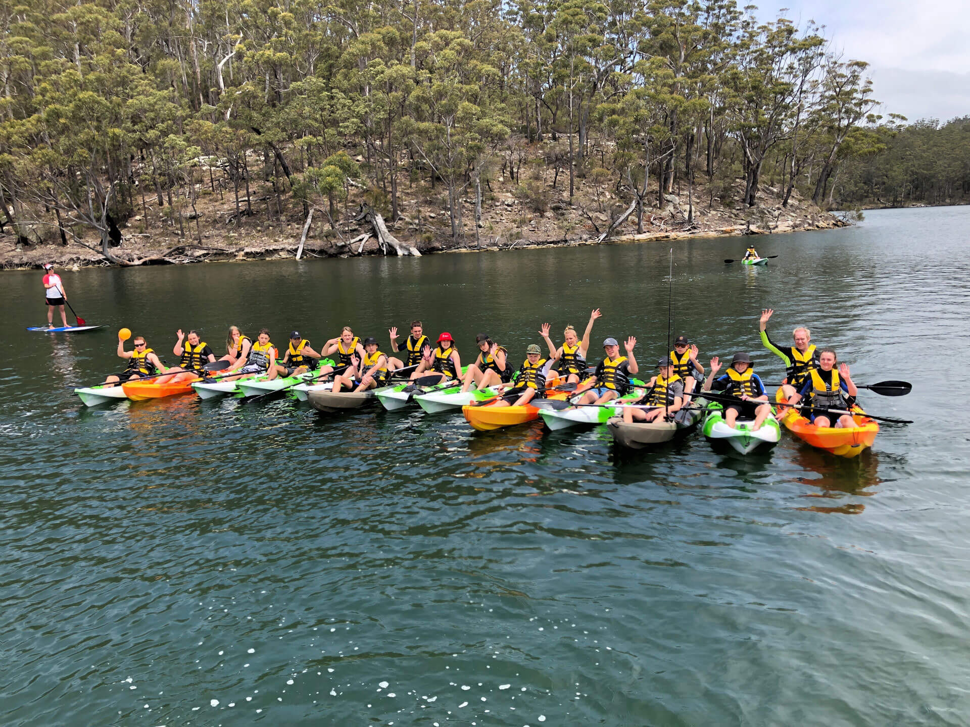 Large group of waving people with life jackets on in a row of green and orange coloured kayaks