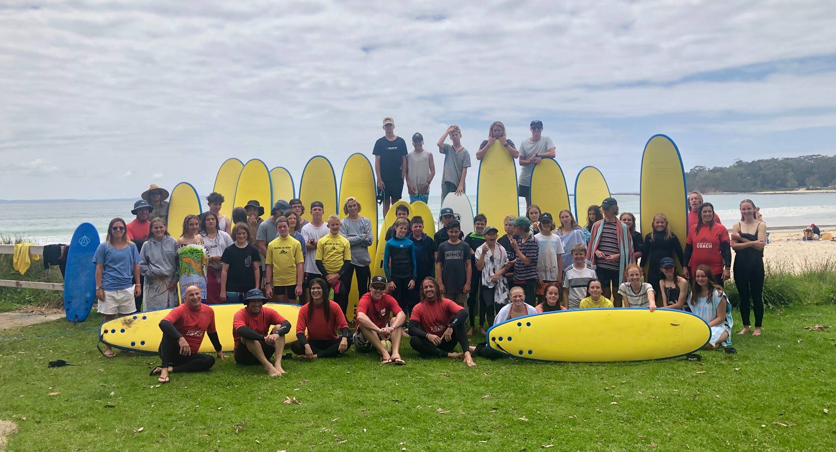 Large group of people with yellow coloured surf boards standing up and laying down on the grass with the ocean in the background