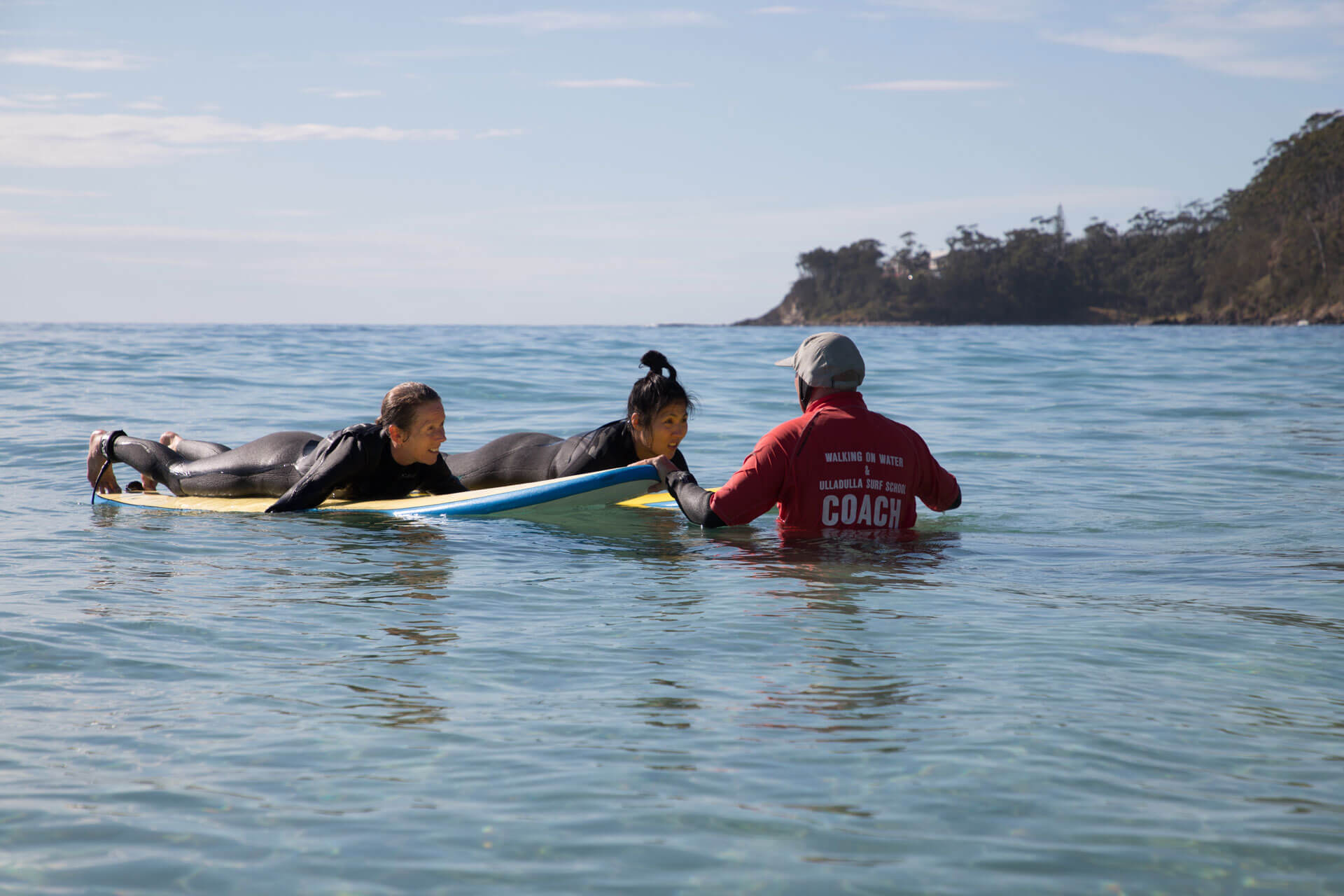 Two women in wetsuits laying down on surf board with their coach during surfing lessons