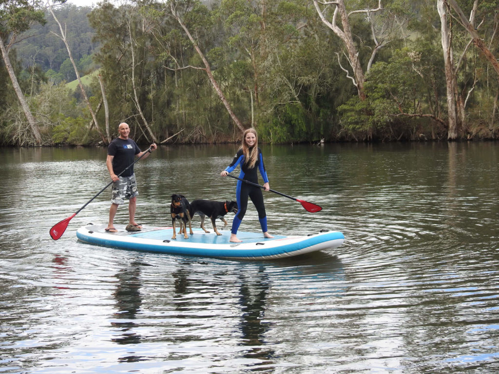 Smiling Coach and girl in wetsuit with two dogs on monster paddle board paddling on the water