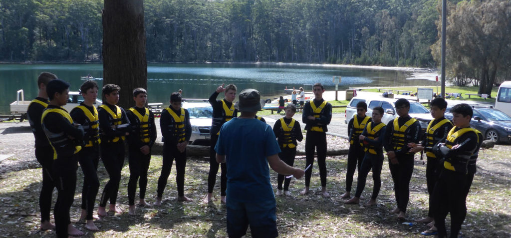Group of young men in matching wetsuits and lifejackets listening to coach talk on surf lessons and water safety