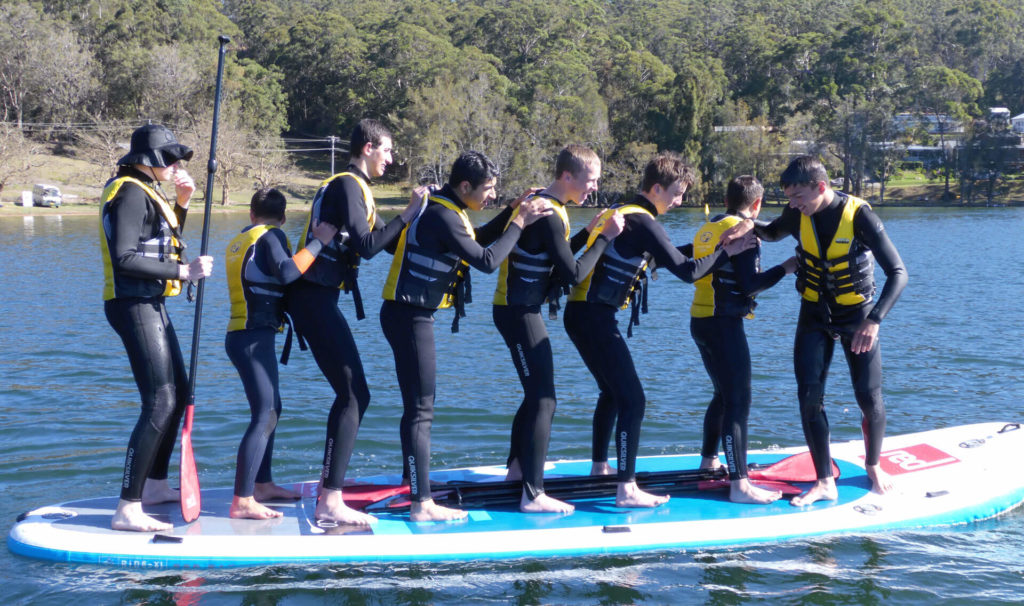 Group of smiling teenage boys in matching wetsuits and life jackets in a row on moster stand up paddle board on the water
