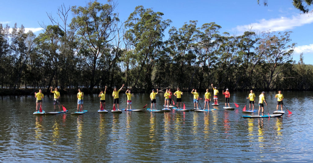 Group of people standing on paddle boards in a row on the water with tree lined bank in the background
