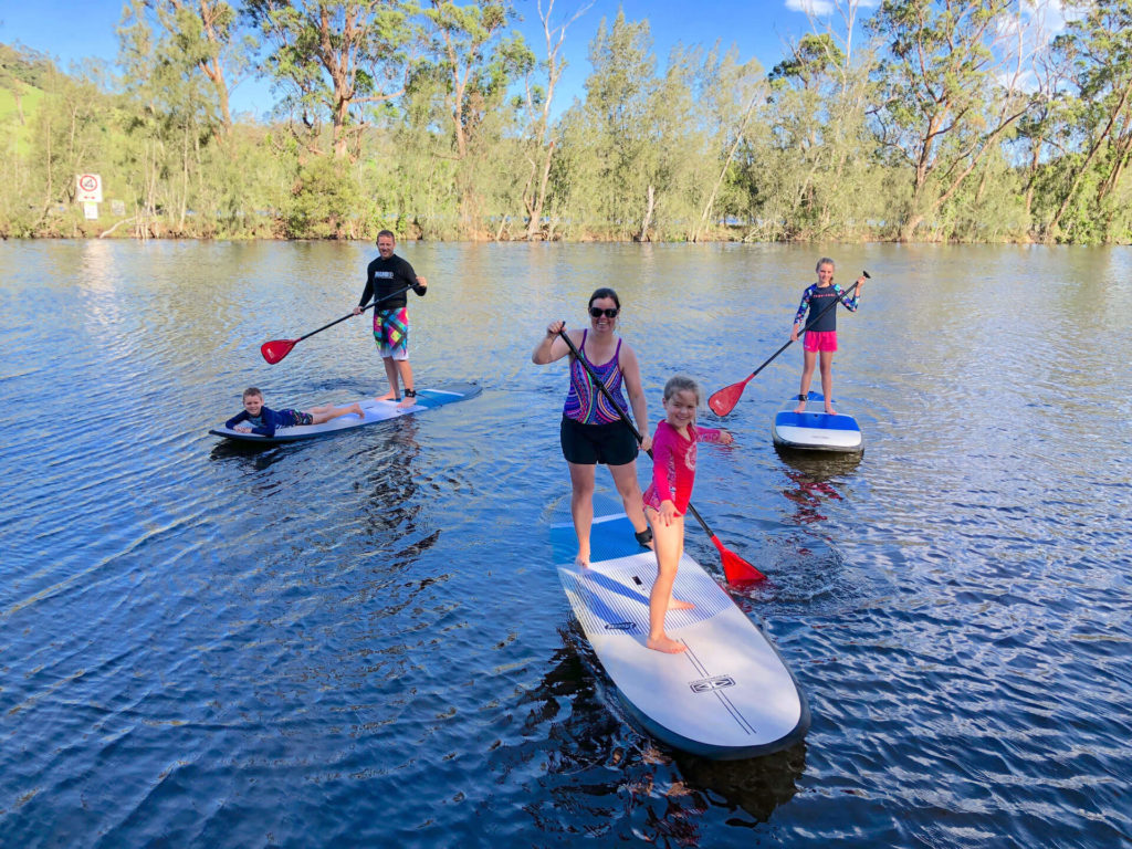 Smiling family standing on paddle boards on the water with tree lined bank in the background