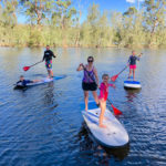Smiling Family Standing On Paddle Boards On The Water With Tree Lined Bank In The Background