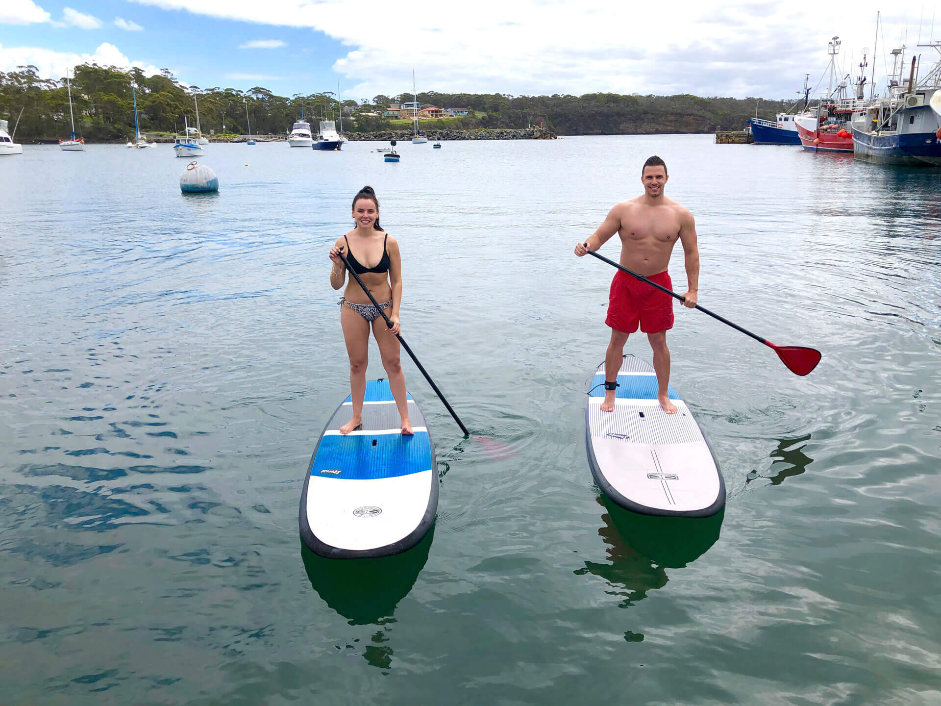 Smiling couple standing on individual paddle boards on the water with boats in the back ground