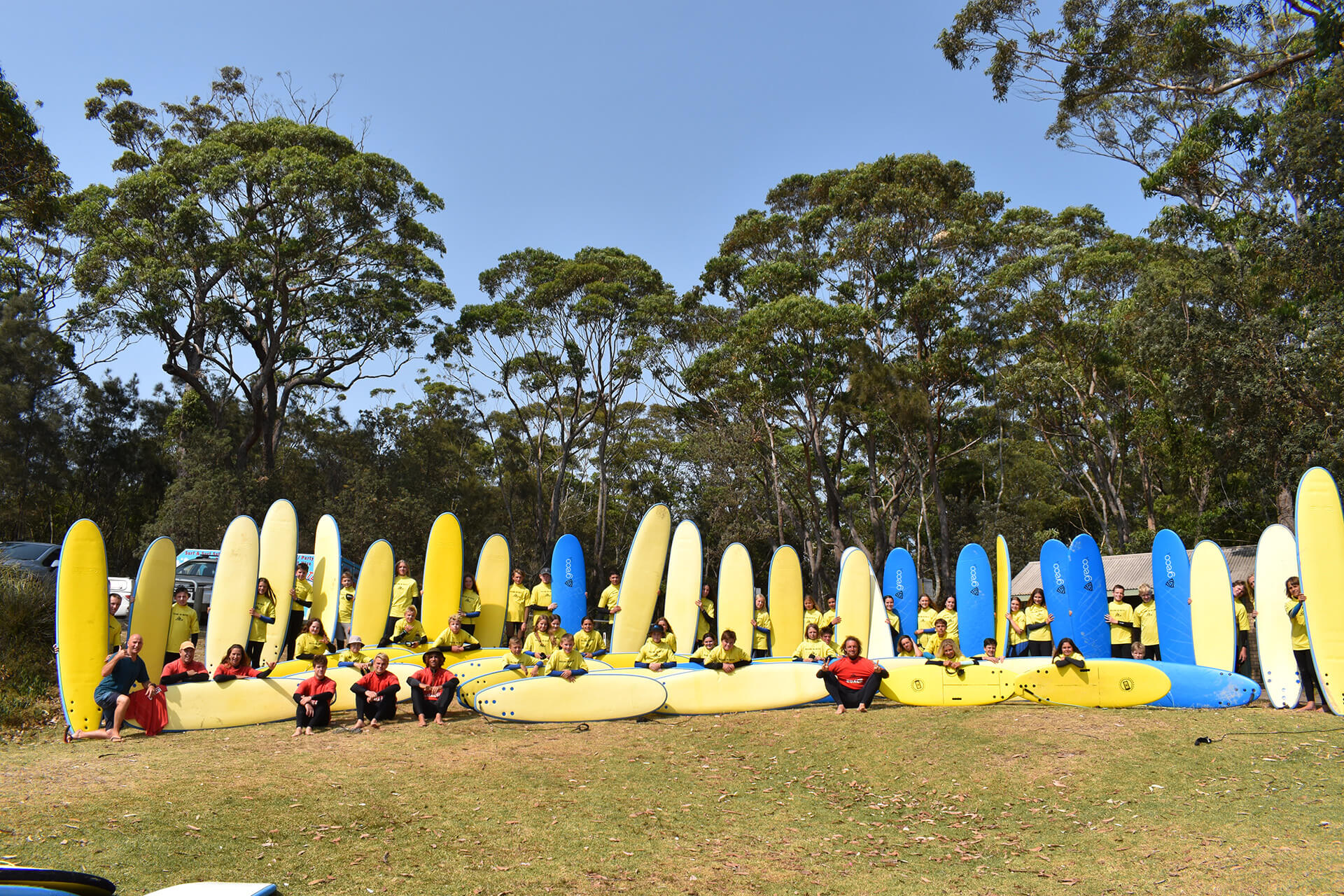 Large group of people with yellow and blue coloured surf boards standing up and laying down on the grass